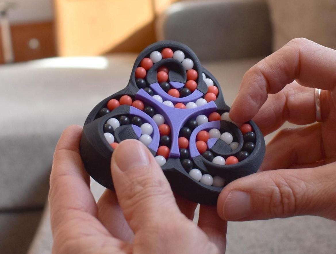 Compact black mechanical puzzle with purple frame and movable red, white, and gray beads, held as a brainteaser fidget toy. - andEleven - The Triad