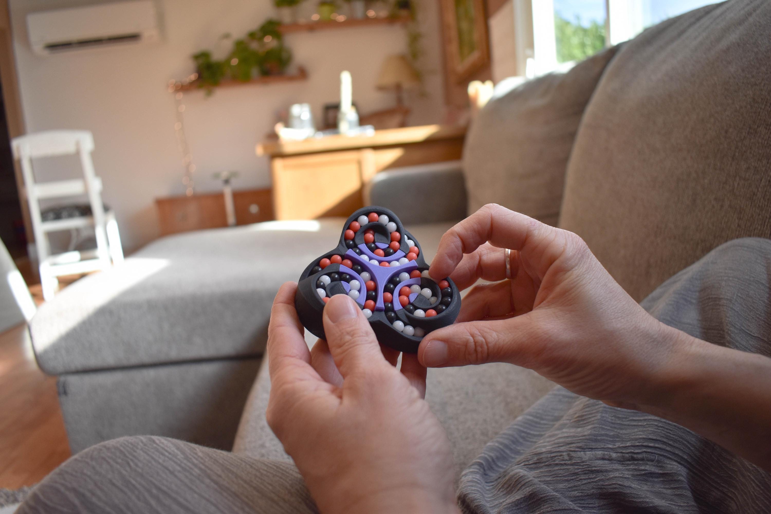 Hands holding a black and red mechanical puzzle brainteaser fidget in a cozy living room setting. - andEleven - The Triad