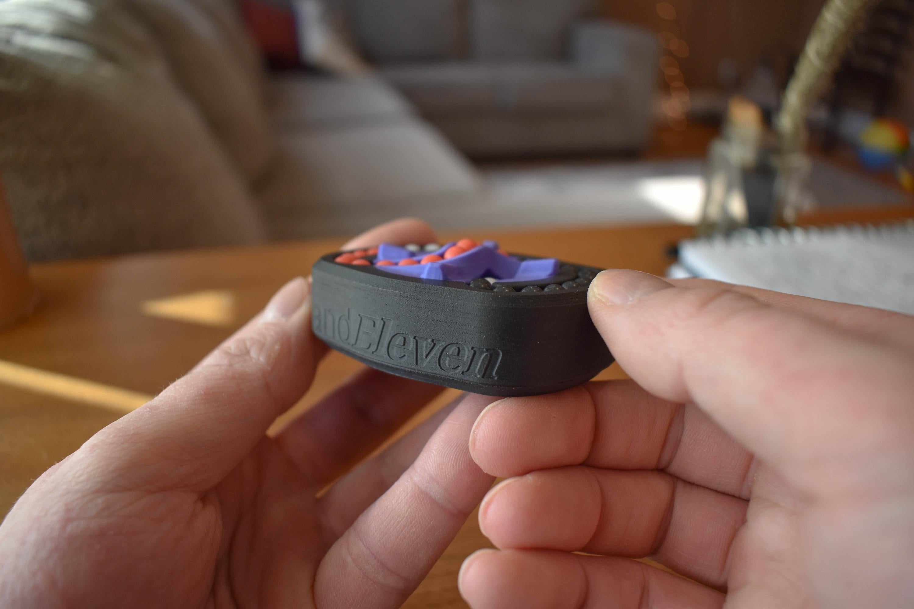 Compact black mechanical puzzle fidget with colorful purple and orange pieces held in hands on couch background, brainteaser . - andEleven - The Infinity (3-Color)