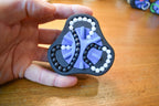 Two-color mechanical puzzle brainteaser fidget with black and purple segments and white beads in hand. - andEleven - The Delta (2-Color)