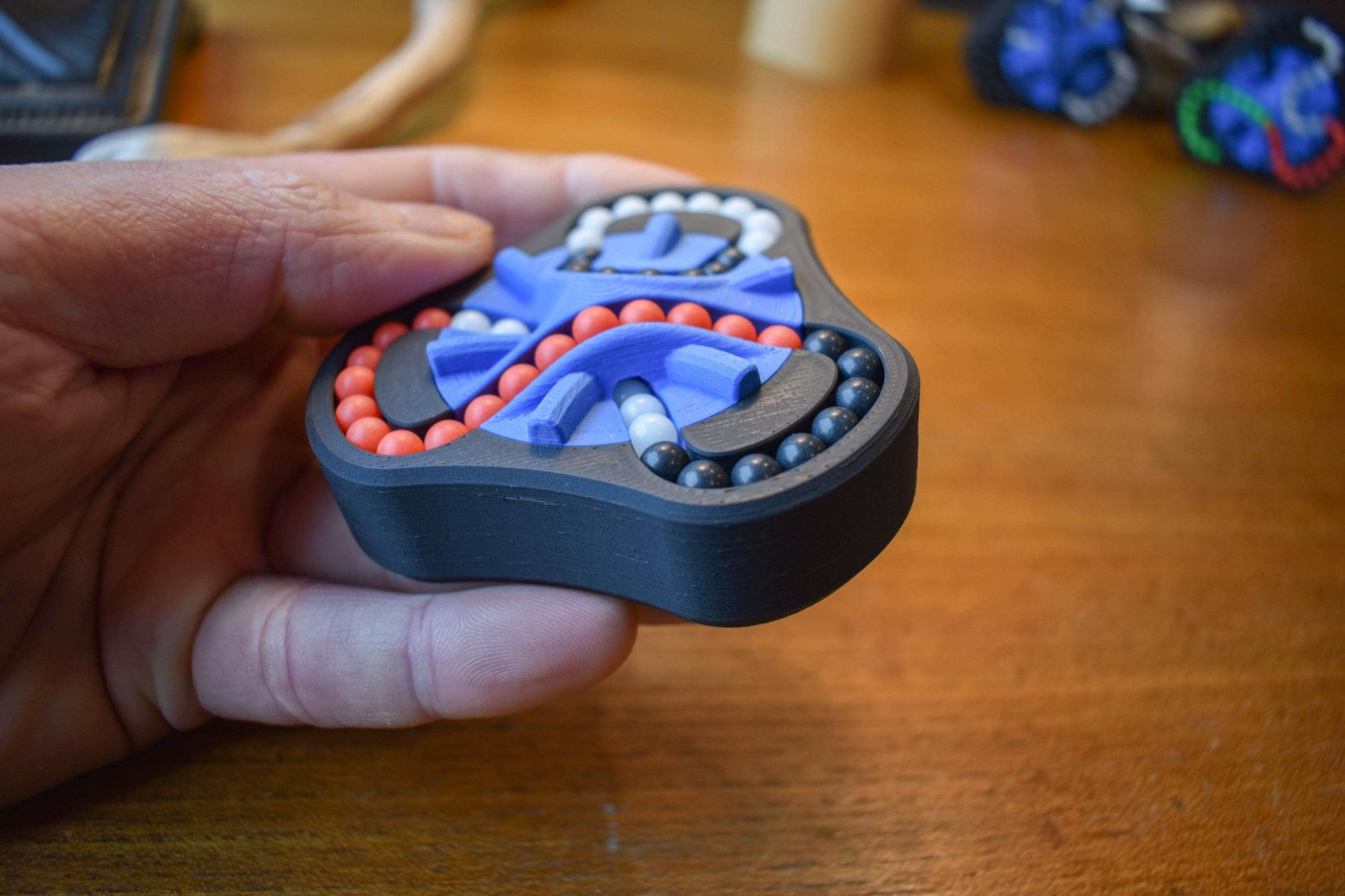 Hand holding The Delta 3-color mechanical puzzle brainteaser with red, blue, and gray beads on wooden surface. - andEleven - The Delta (3-Color)