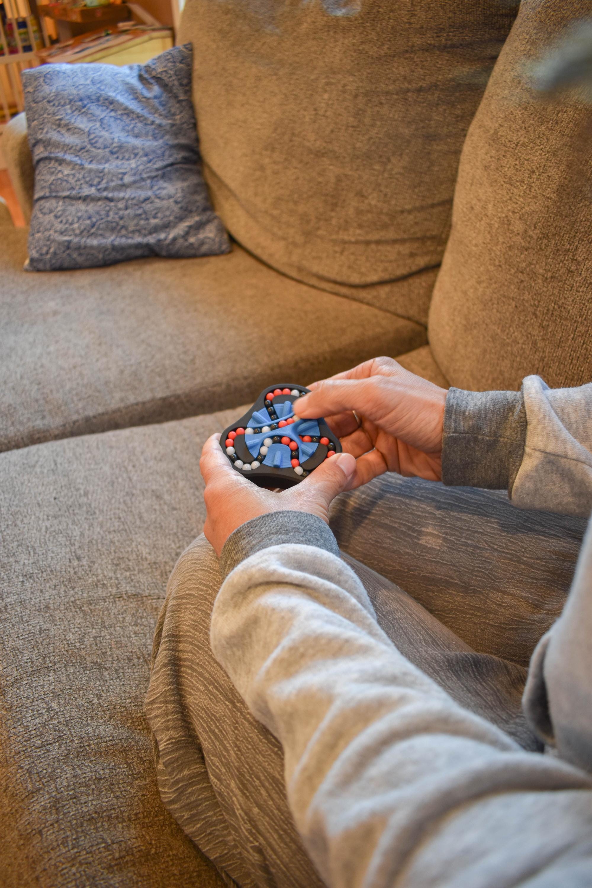 Hands holding a red, black, and blue mechanical puzzle brainteaser fidget on a beige couch. - andEleven - The Delta (3-Color)